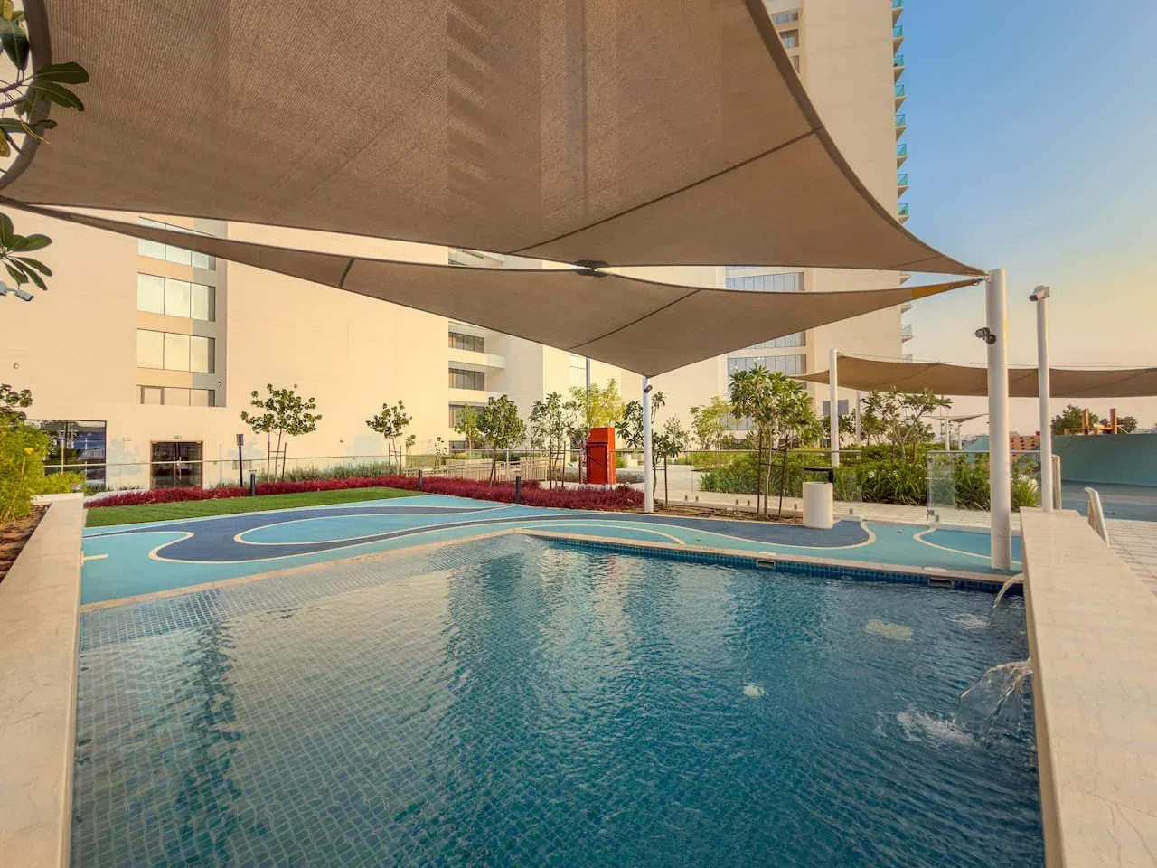 Una piscina al aire libre está bajo grandes velas de sombra junto a un patio ajardinado.