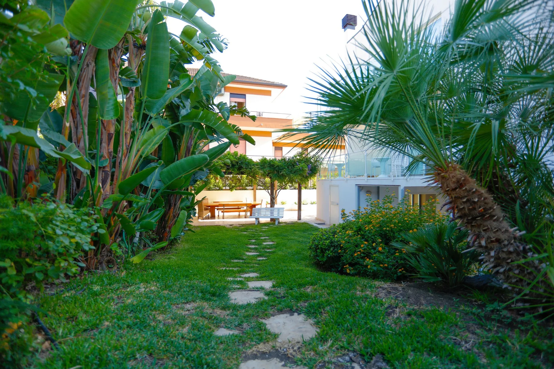 A garden path leads through tropical plants toward a seating area and building.
