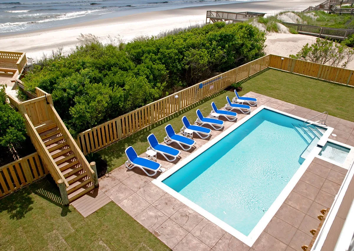 An outdoor swimming pool with blue lounge chairs, beside a sandy beach.