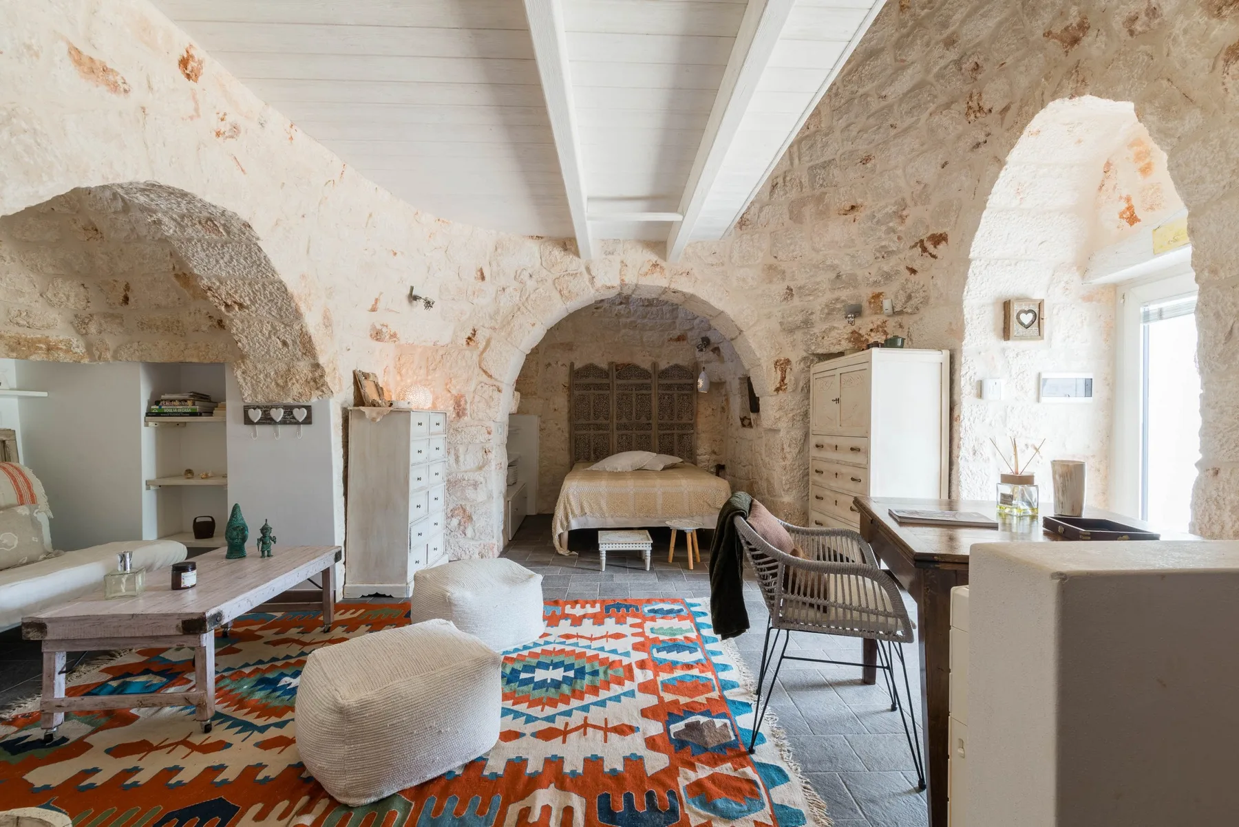 A bright stone bedroom and sitting area with a bed, tables, and patterned rugs.