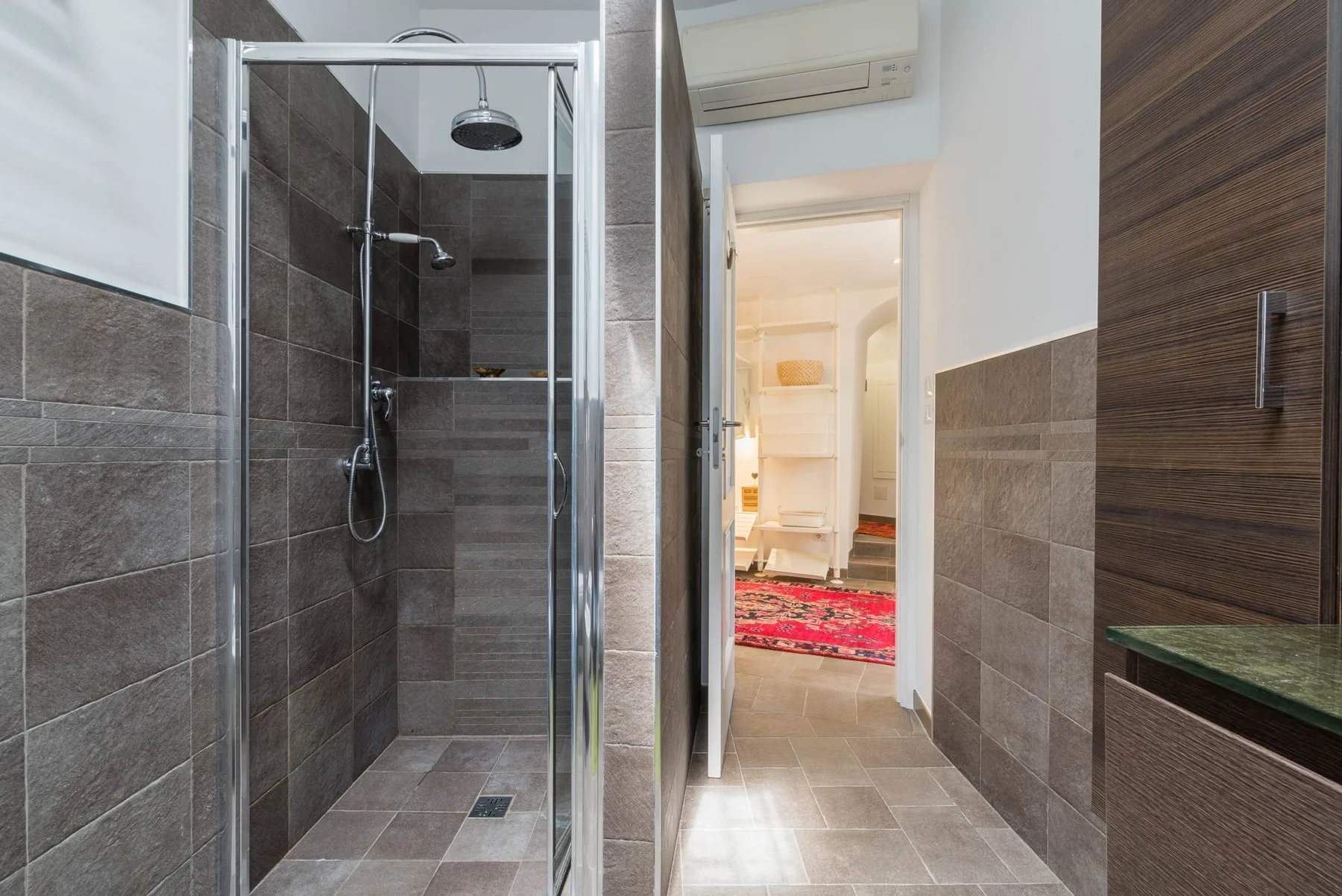 A tiled bathroom with a glass shower and an open doorway to a bright room.