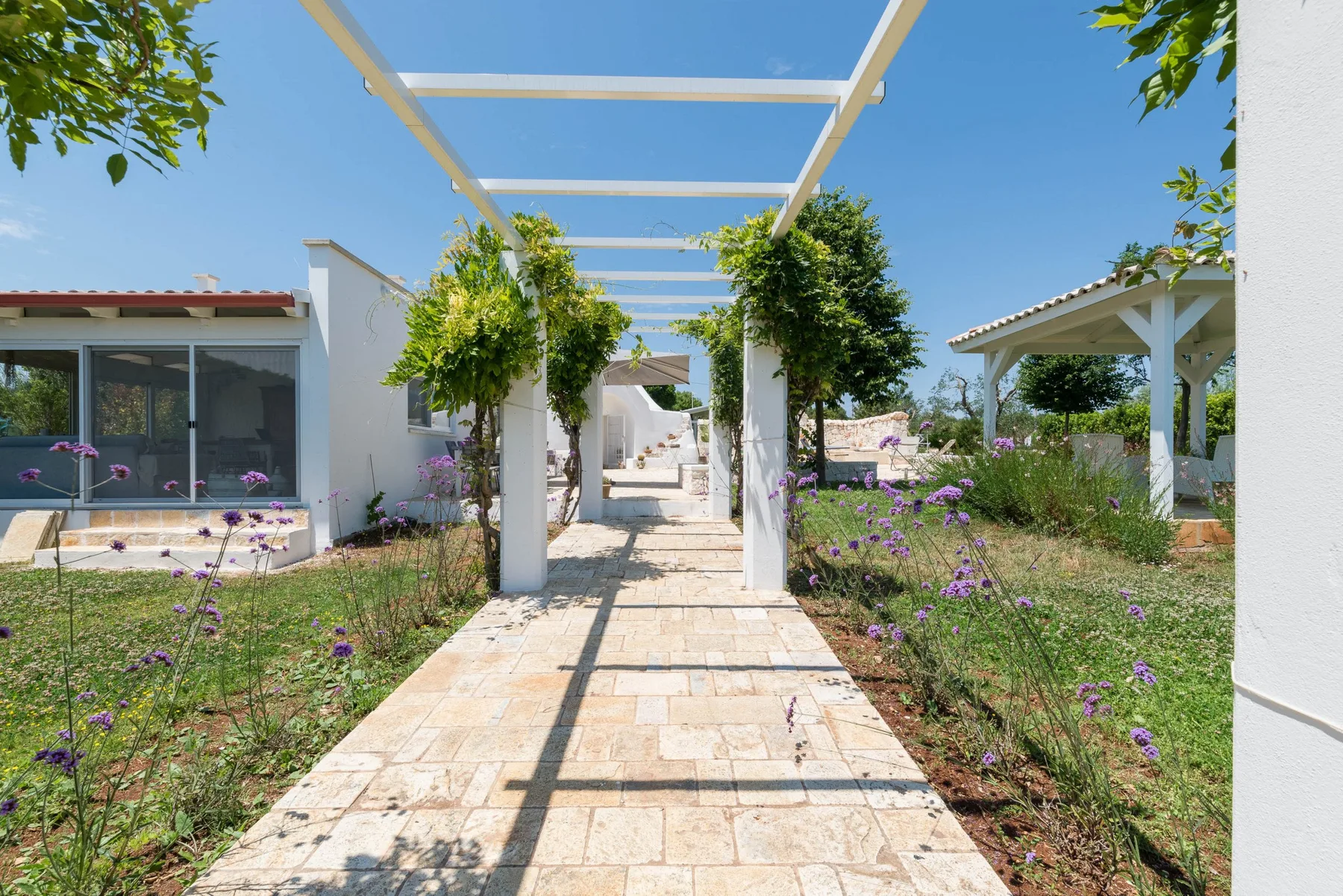 A stone walkway passes through a landscaped garden beneath a white pergola.