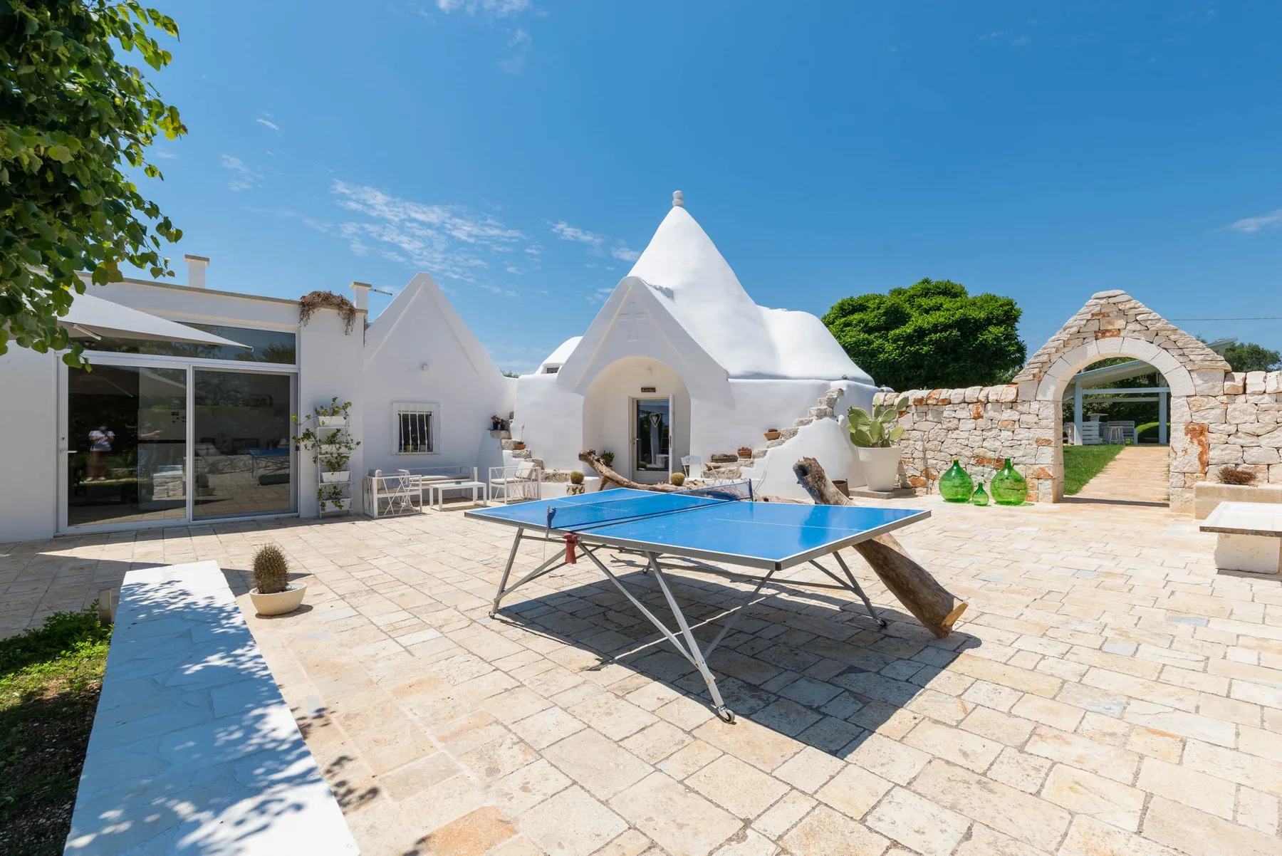A sunny stone courtyard with a blue table tennis table and white buildings.