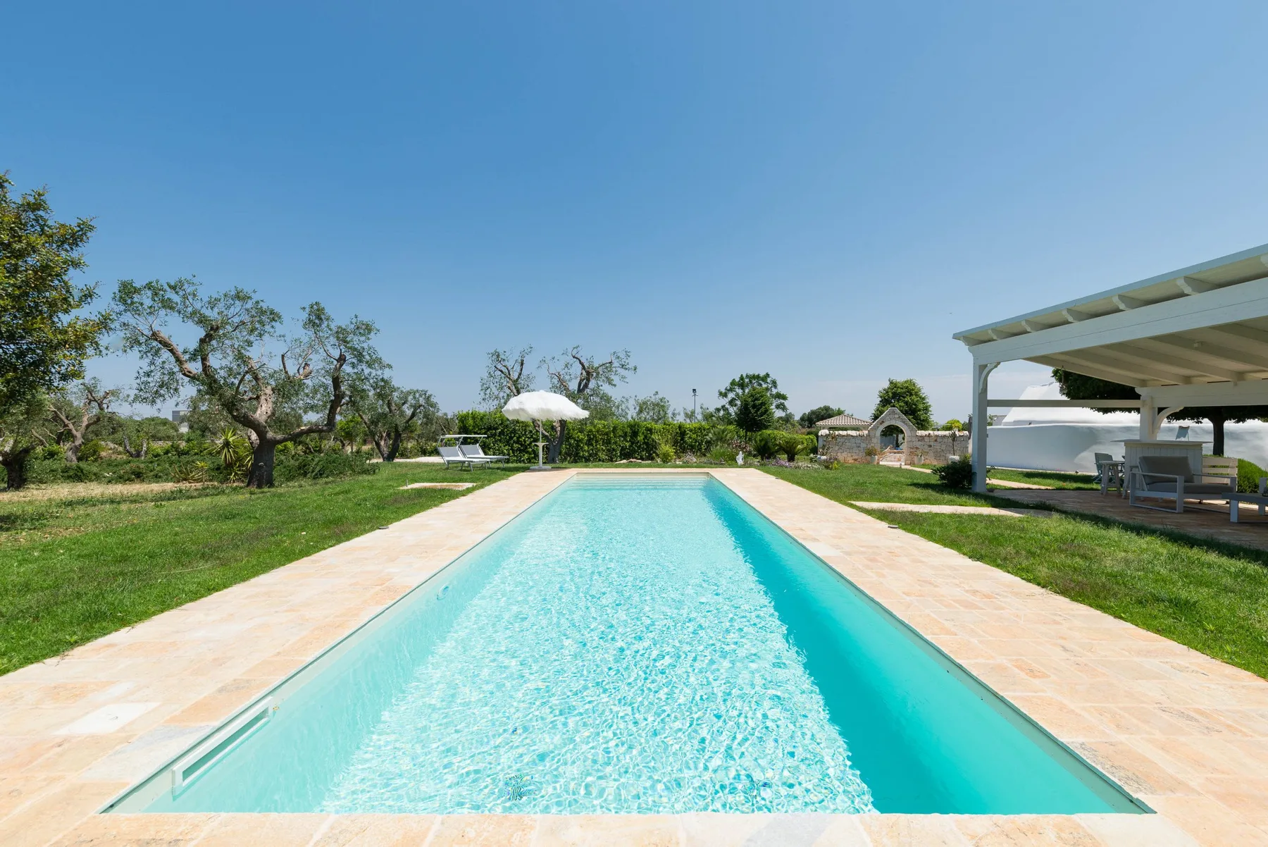 A swimming pool bordered by a pale stone deck sits in a green garden.