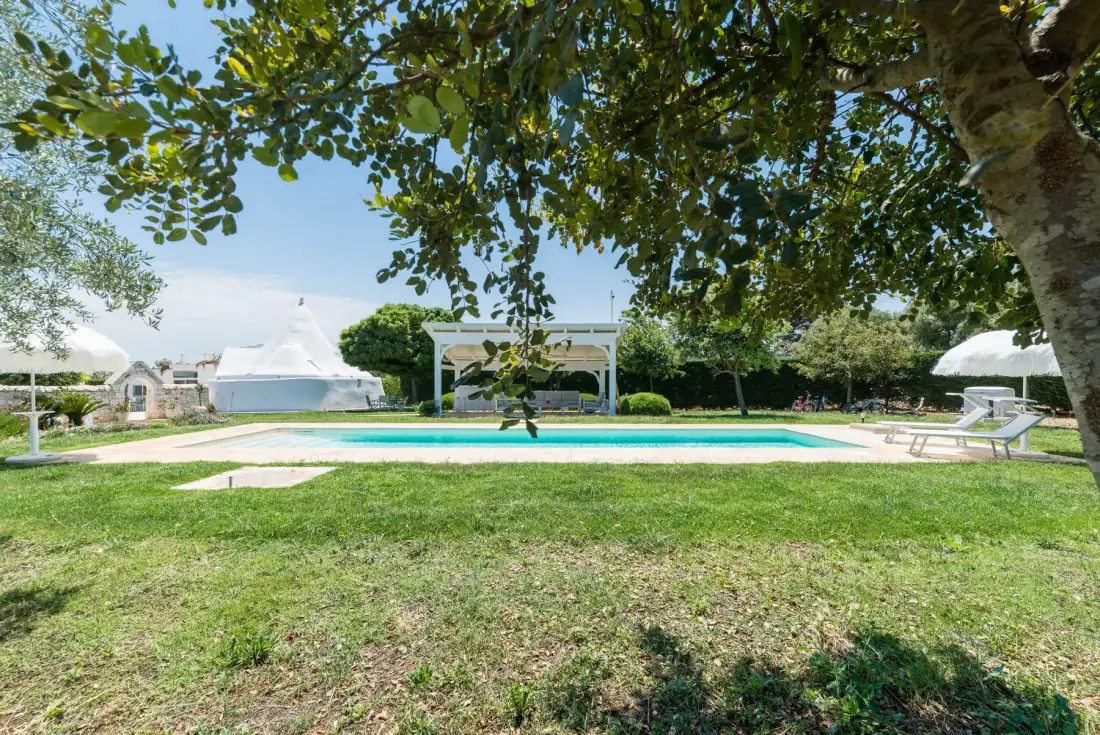 An outdoor swimming pool sits beside a grassy lawn and shaded seating area.