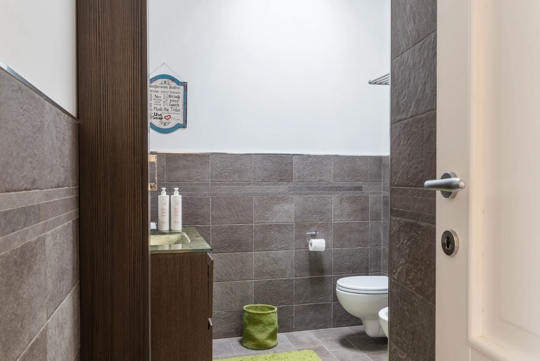 A tiled bathroom with a toilet, sink cabinet, and green floor mat.