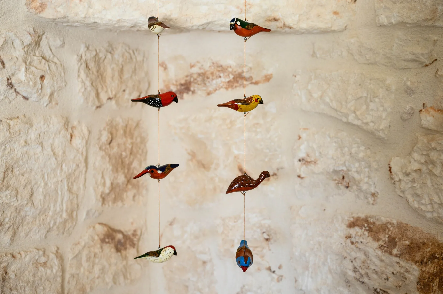 Small colorful bird ornaments hang against a rough stone wall.