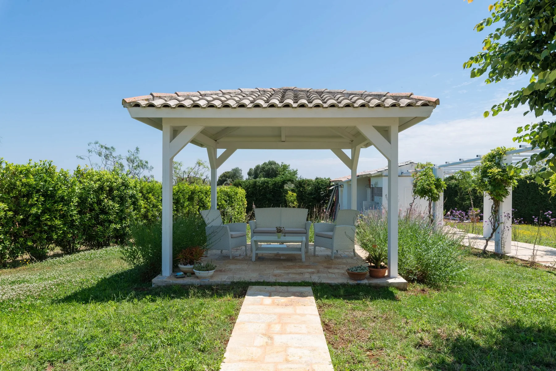 A garden gazebo with seating stands beside a paved path and green hedges.