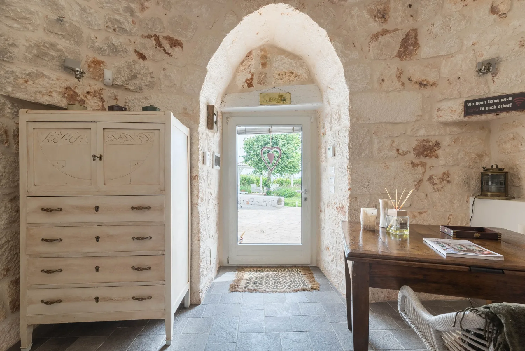 A rustic stone room with a central arched doorway and a desk beside the window.