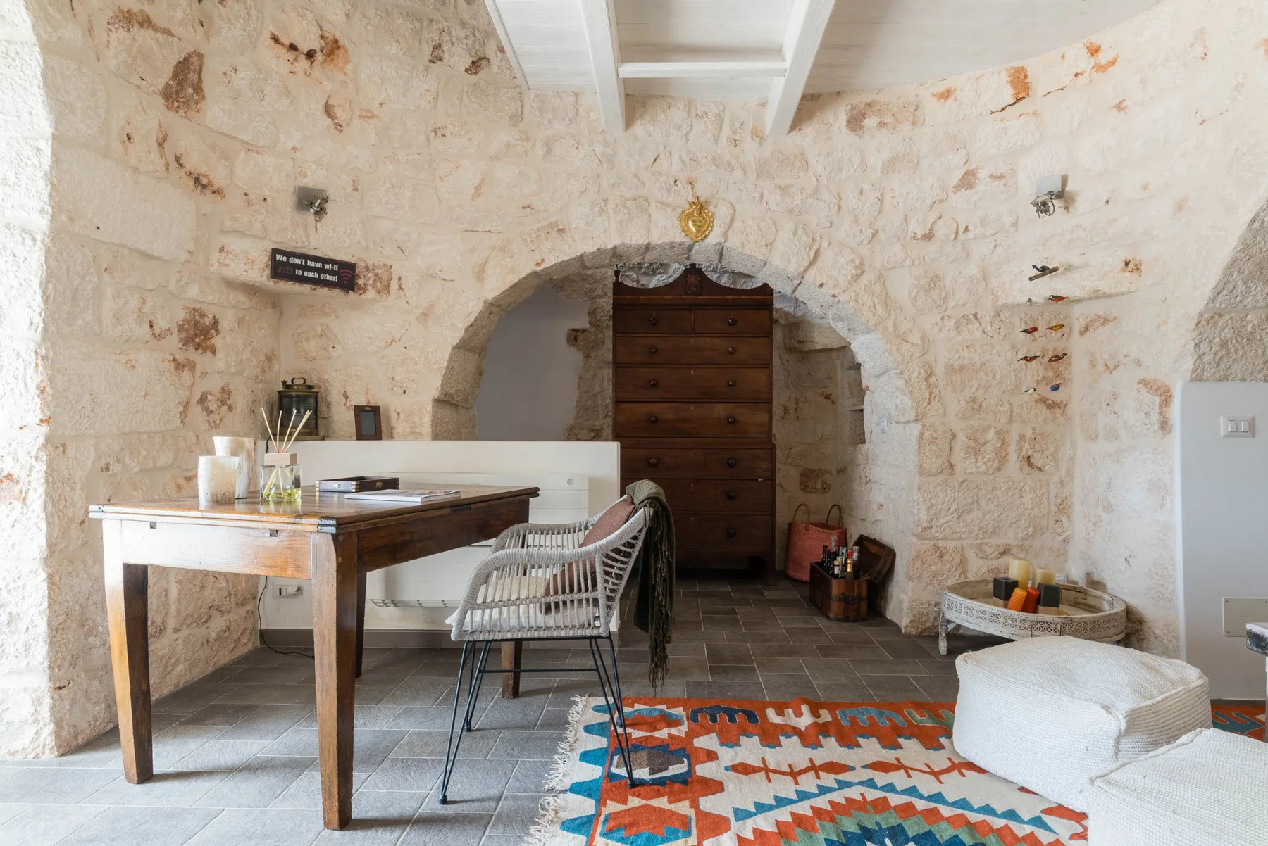 A rustic stone room with a wooden table, a central arched doorway, and a patterned rug.