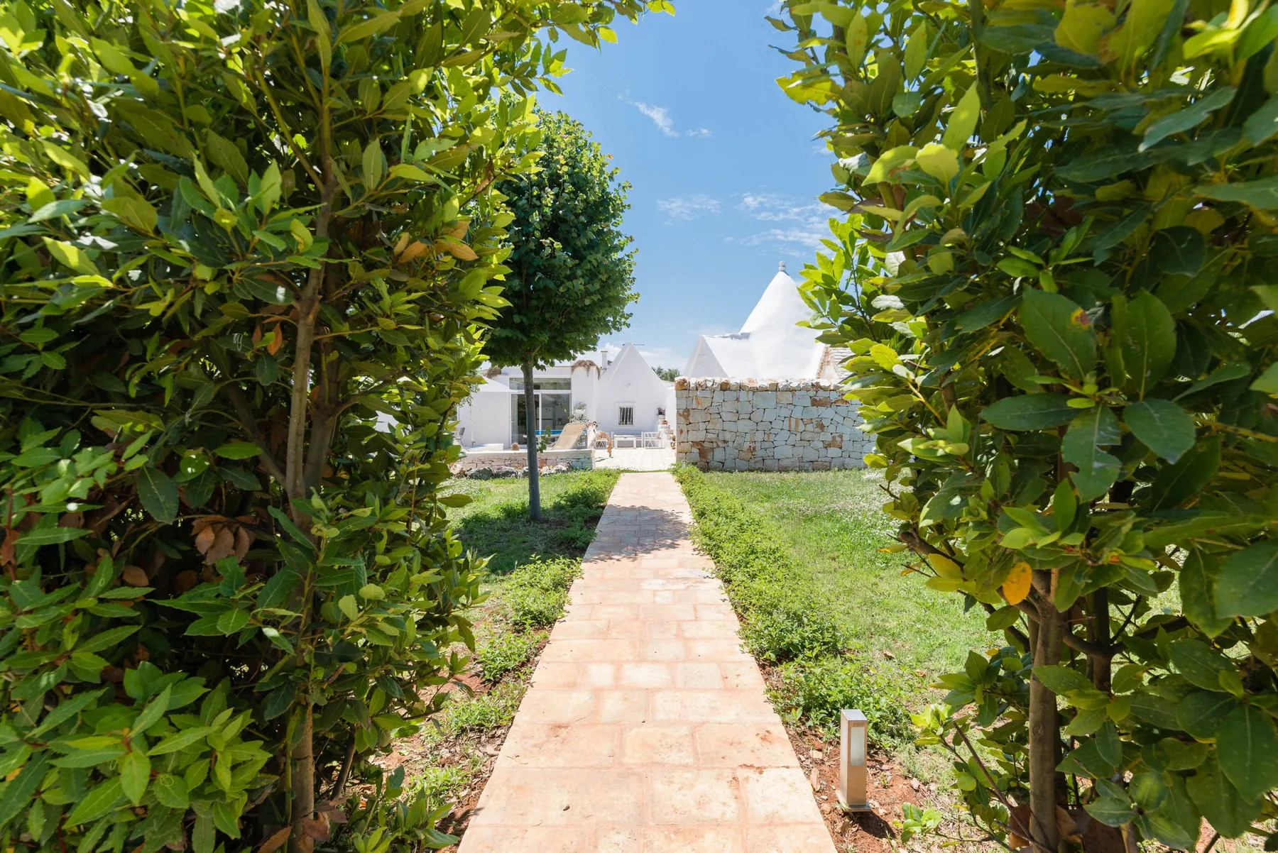 A stone walkway passes between leafy trees toward white buildings beneath a clear sky.