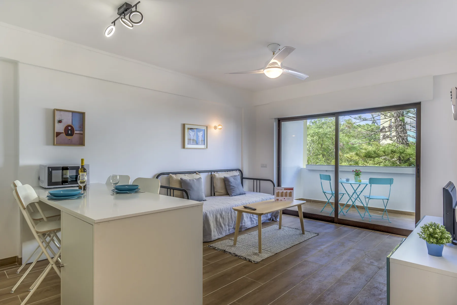 A bright studio living area with a dining counter and a balcony overlooking trees.