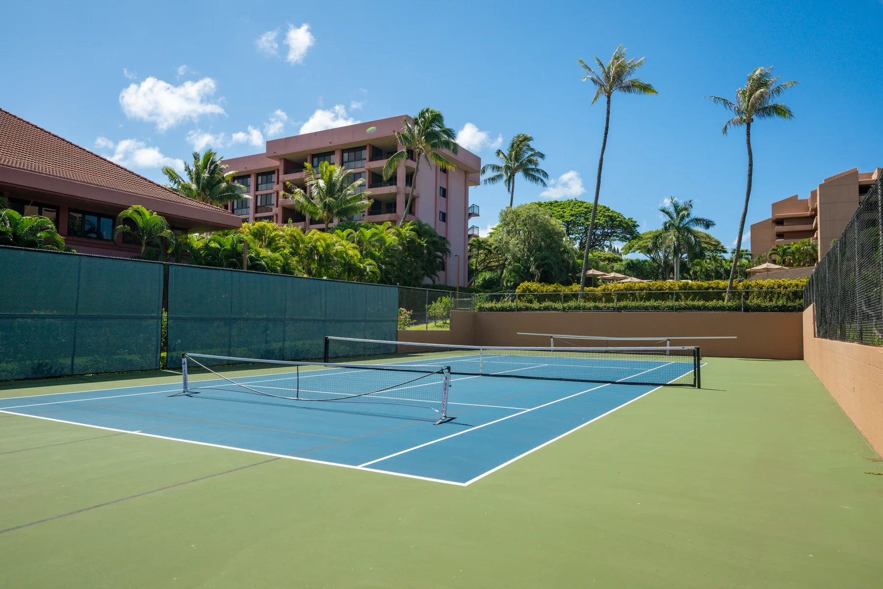 An outdoor tennis court is surrounded by tropical landscaping and buildings.