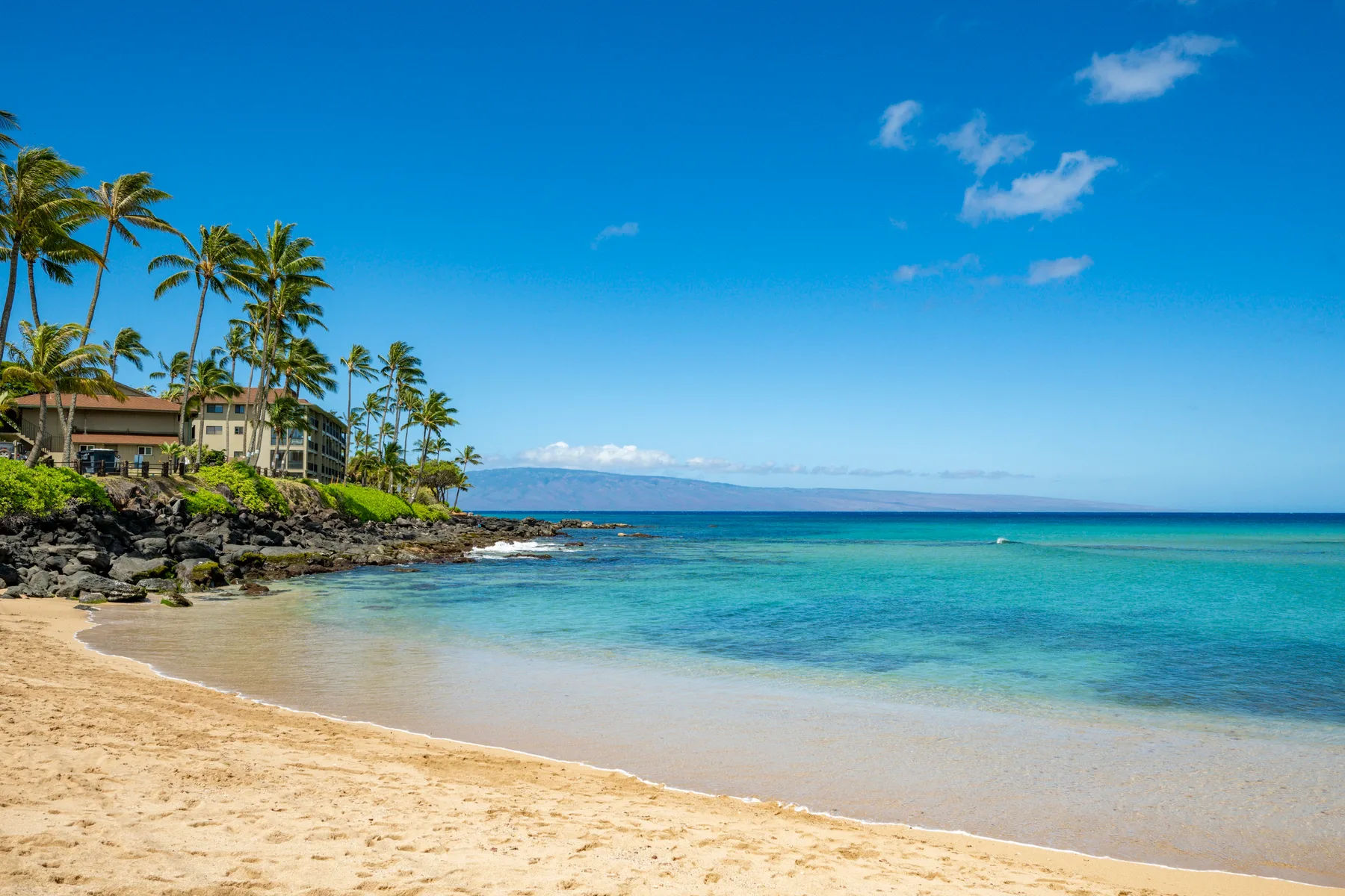 A sandy beach borders clear turquoise water, with palms and buildings along the rocky shore.