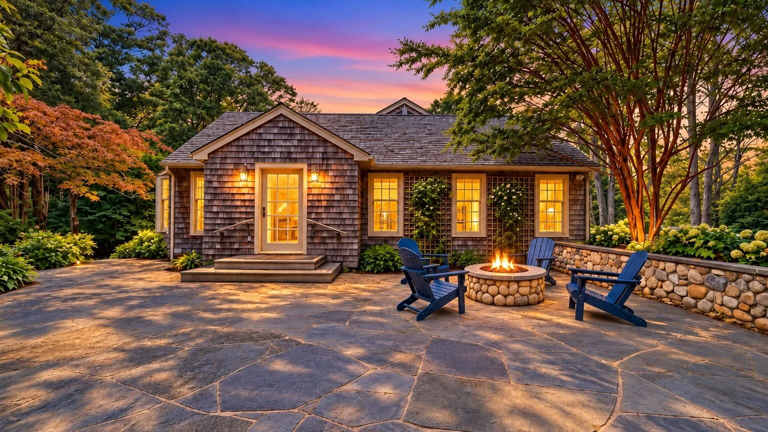 A cottage exterior with a stone patio, Adirondack seating, and a lit fire pit.