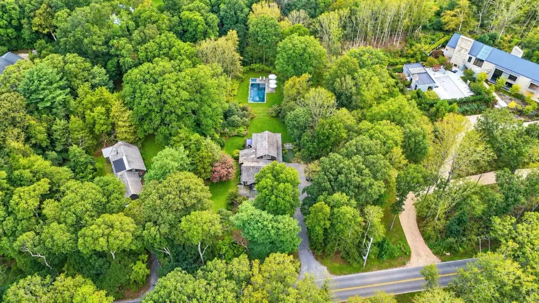 An aerial view of a wooded property with buildings, a pool, and a road.
