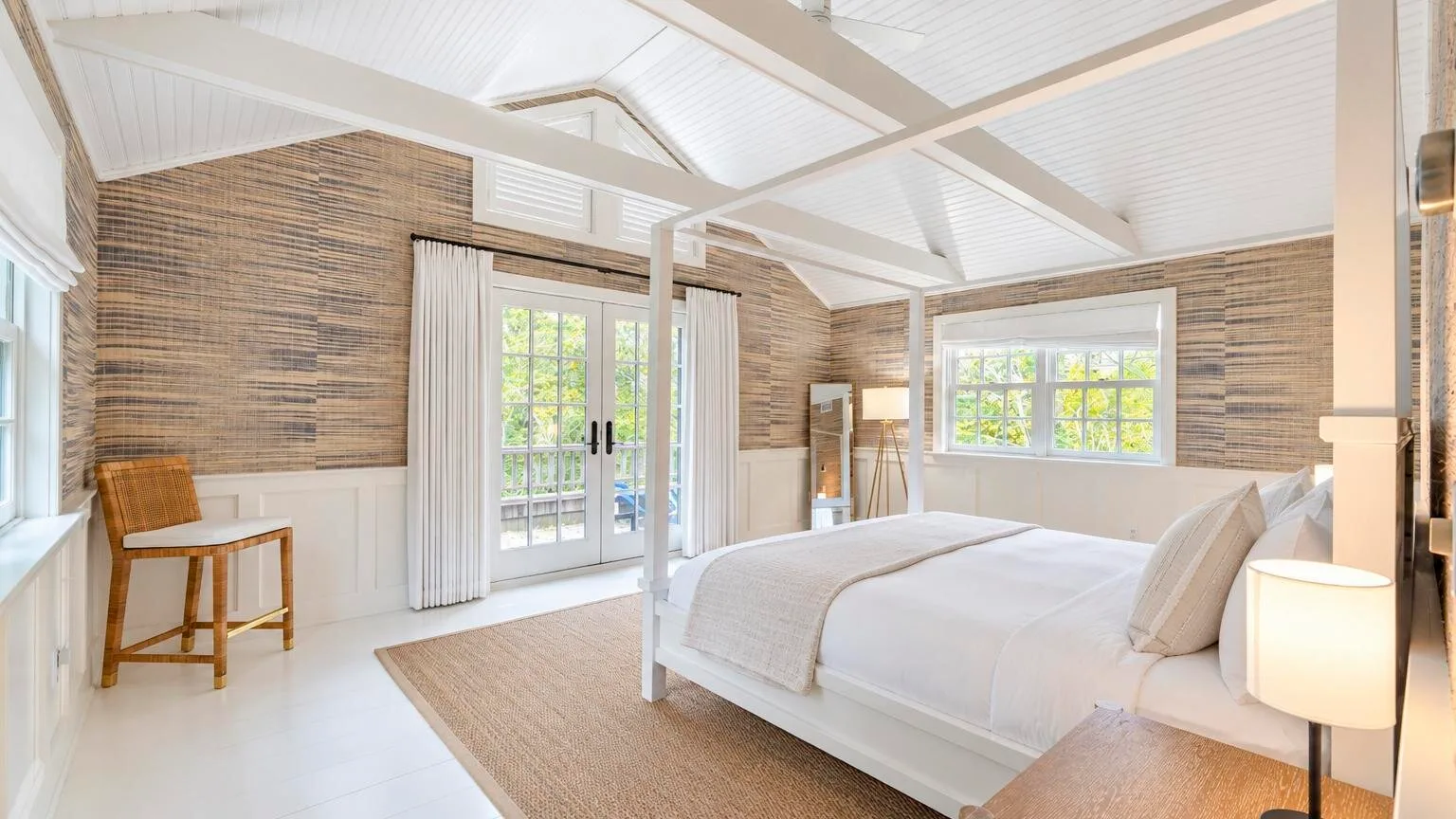A bright bedroom with a large bed, woven wall covering, and windows.