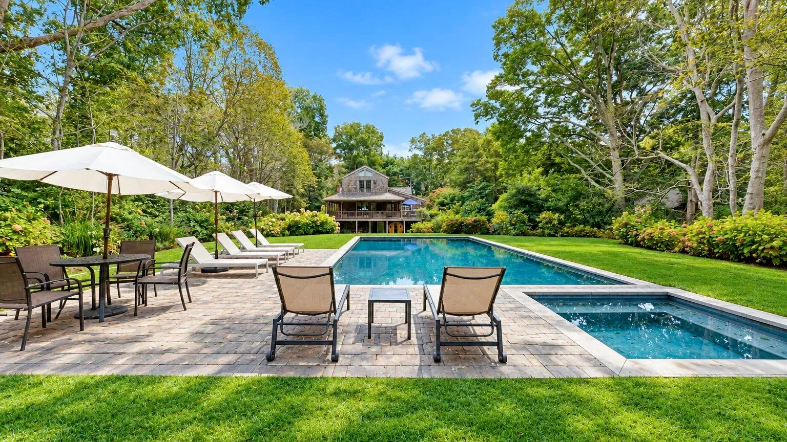 Outdoor pool beside a paved patio with lounge seating and trees.