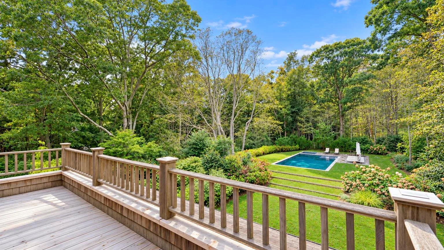 A wooden deck overlooks a green lawn, landscaped garden, and swimming pool.