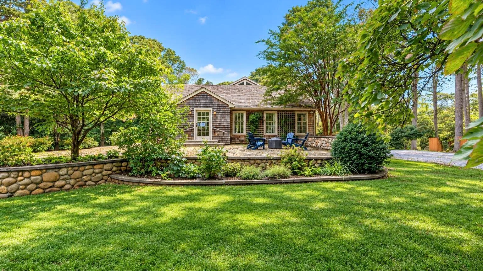 A cottage sits behind a broad lawn, surrounded by leafy trees and shrubs.