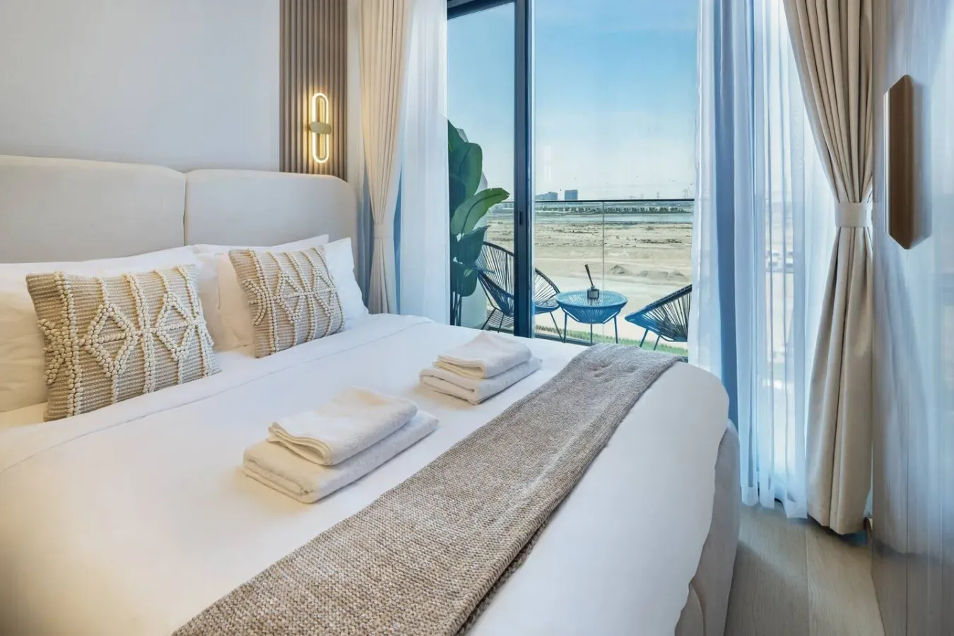 A bright bedroom with a neatly made bed beside a large window overlooking the water.