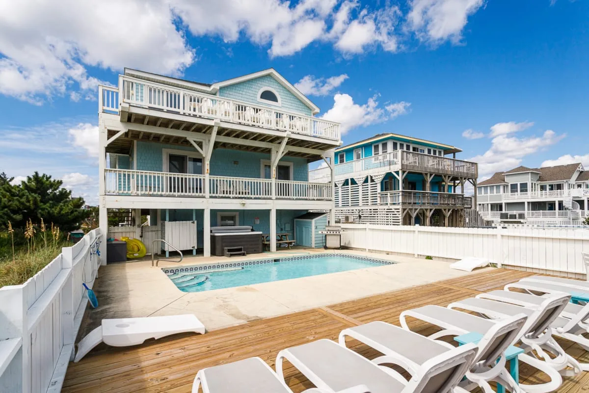 A backyard pool sits beside a multi-level coastal-style house under a blue sky.