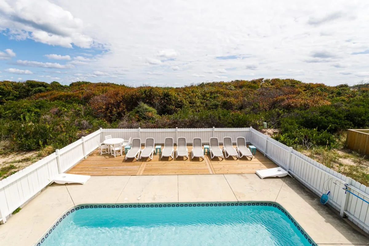 A swimming pool and deck seating overlook a fenced coastal landscape.