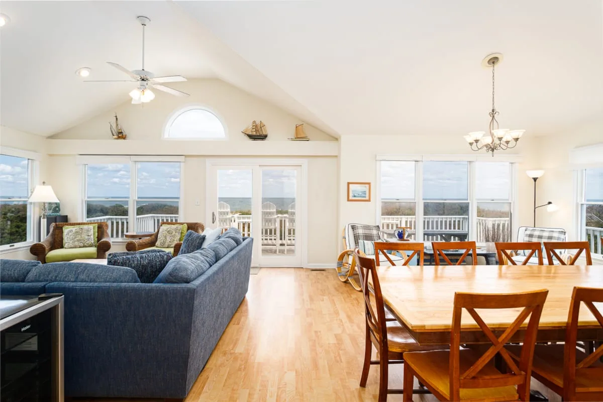 A bright open-plan living and dining area has large windows and a wooden dining table.