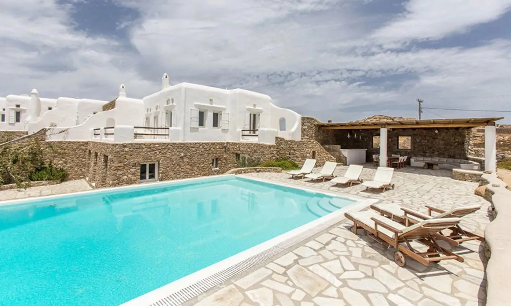 A swimming pool beside white buildings, stone paving, and lounge seating.