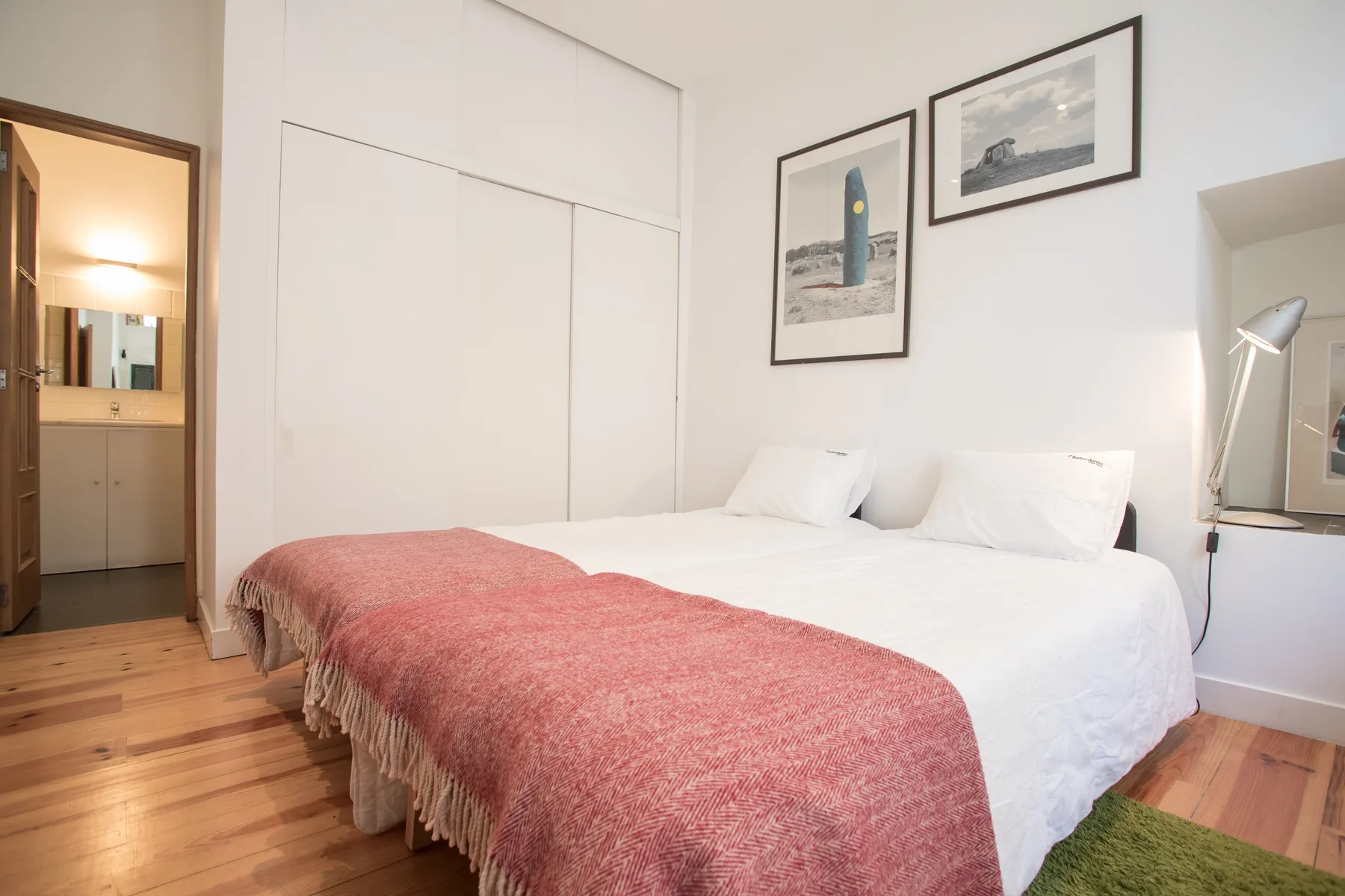 Un dormitorio con una cama blanca, una manta rosa y cuadros en la pared.