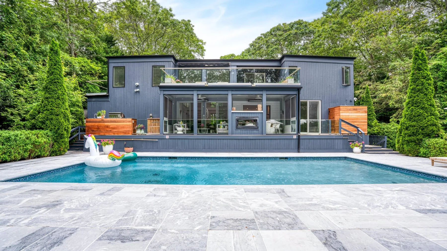 Una casa moderna con piscina y patio pavimentado, rodeada de árboles.