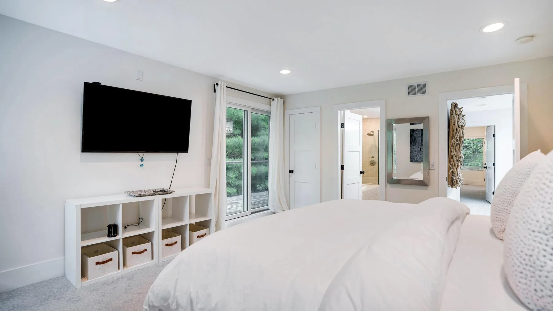 A bright bedroom with a large bed, wall-mounted television, and windows.