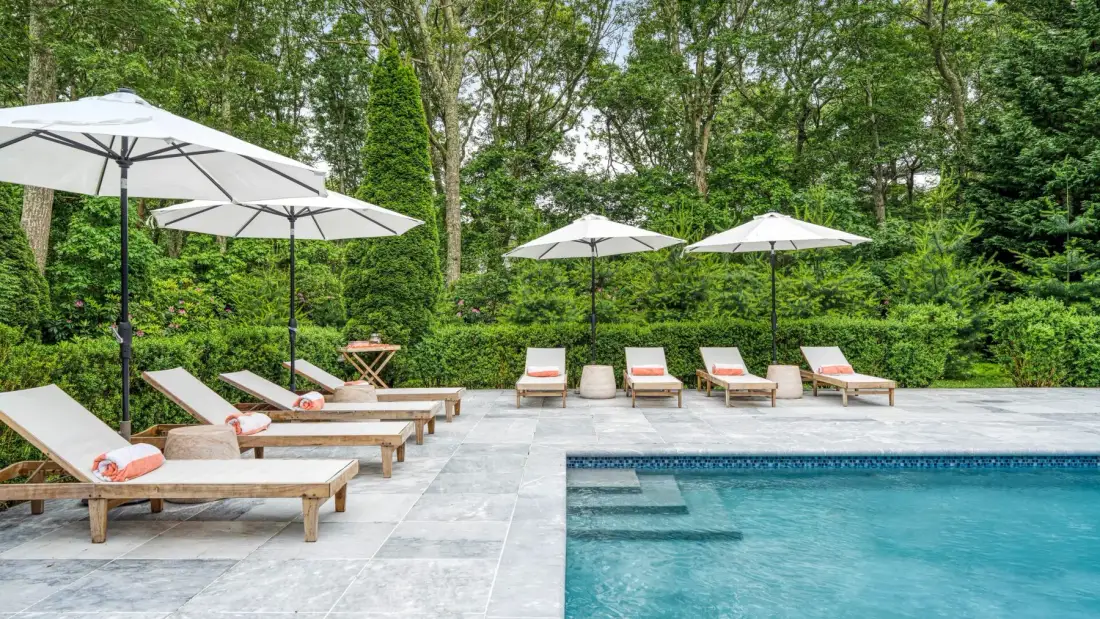 An outdoor swimming pool is bordered by a stone patio with lounge seating and umbrellas.