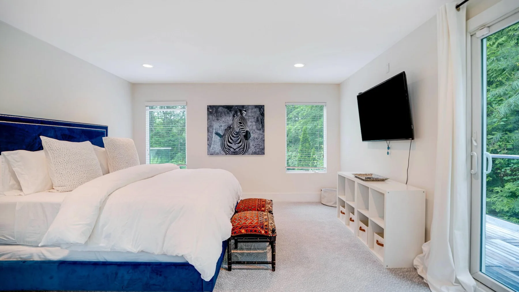 A bright bedroom with a blue bed, wall-mounted television, and windows.