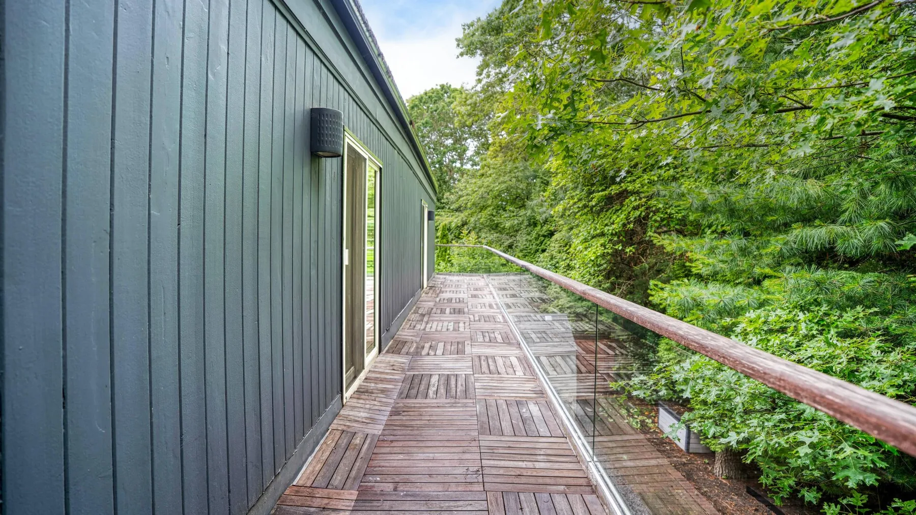 An outdoor wooden deck runs alongside a dark-sided building beneath leafy trees.