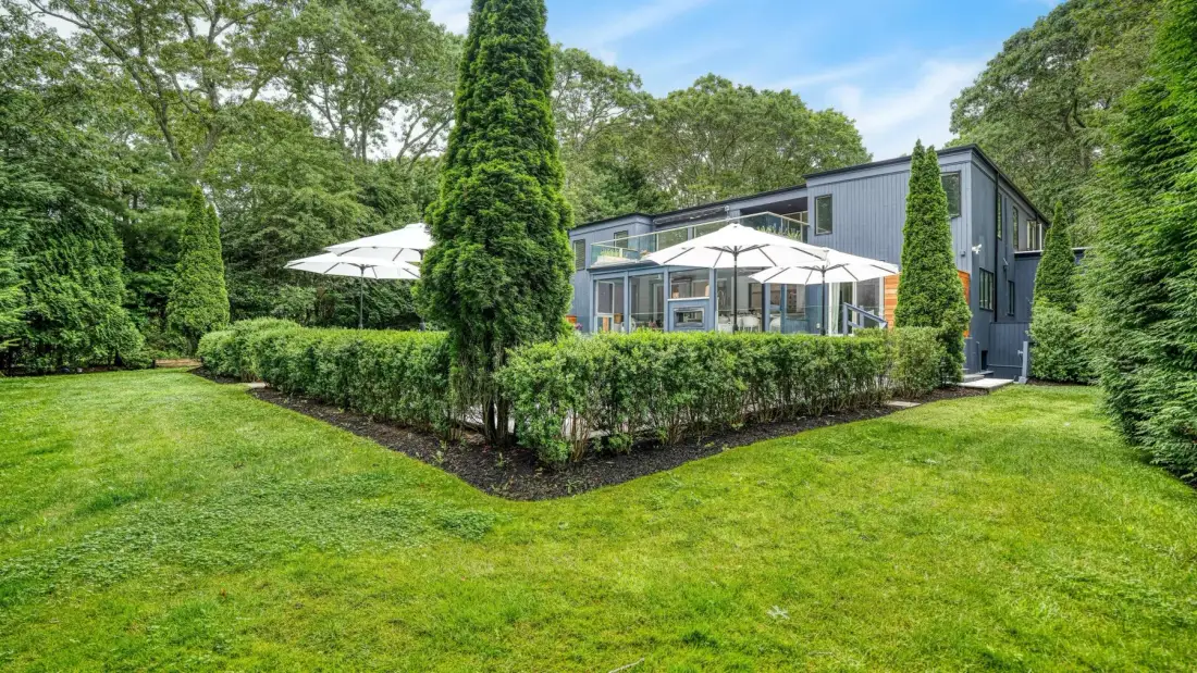 A modern house stands behind a hedge, with a lawn and patio umbrellas.