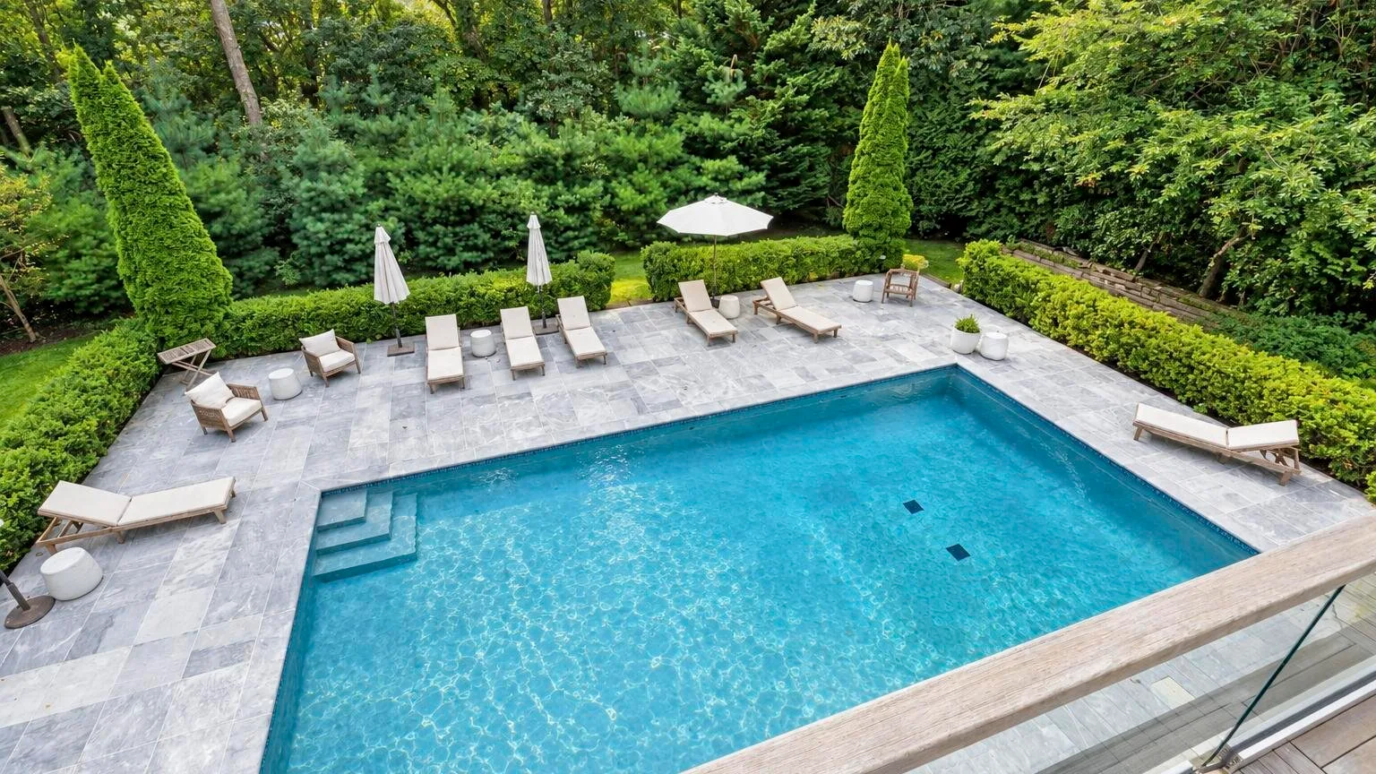 An outdoor swimming pool sits beside a paved patio surrounded by greenery.