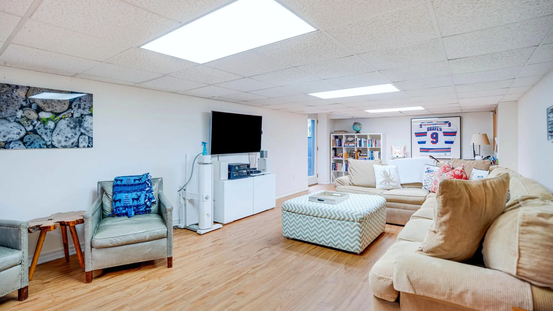 A basement living room with cushioned seating, a television, and a patterned ottoman.