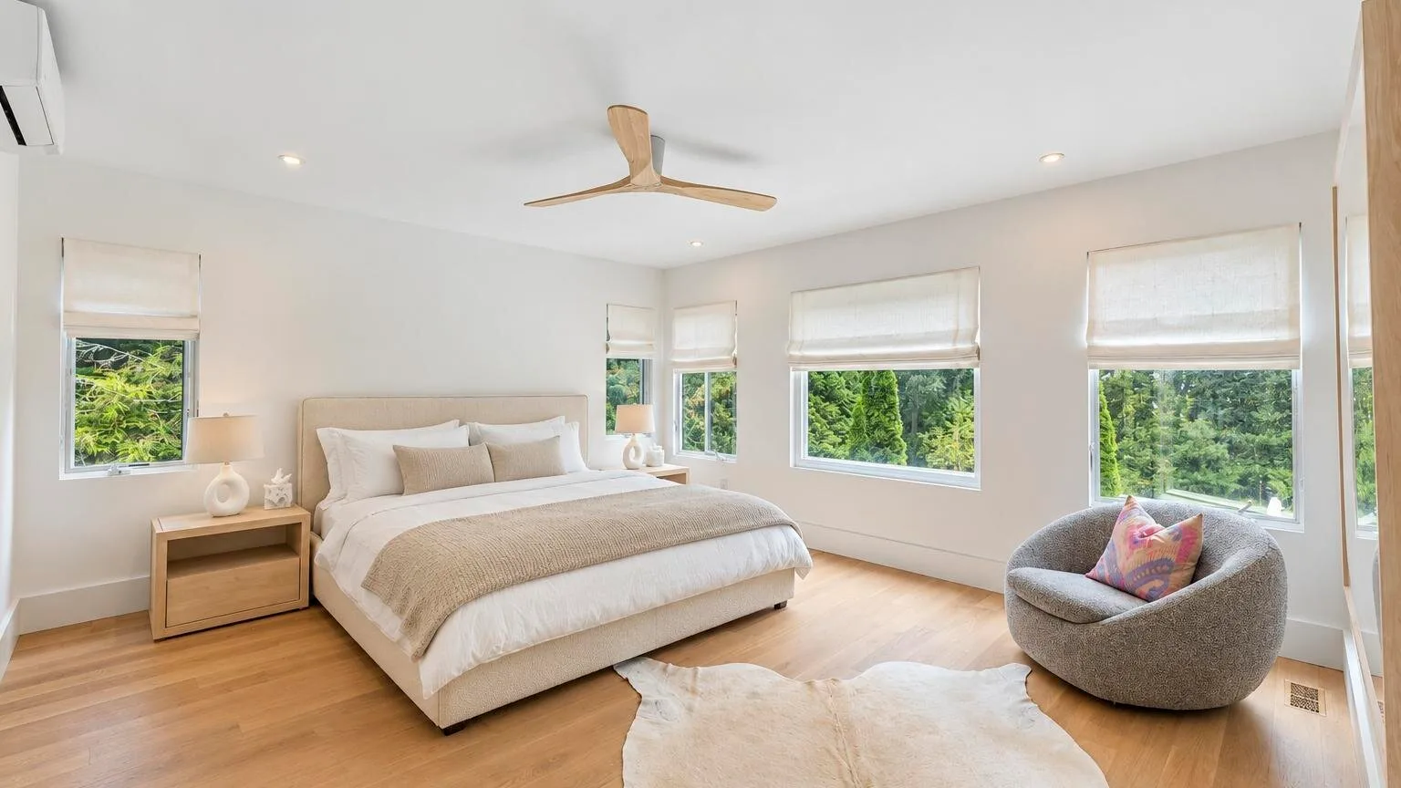 A bright bedroom with a large bed, a rounded chair, and windows overlooking greenery.