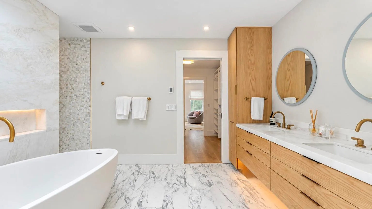 A bright bathroom with a freestanding bathtub, wood vanity, and round mirrors.