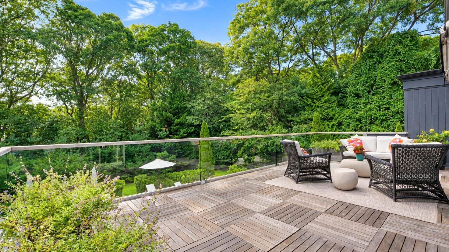 A wooden outdoor deck with seating overlooks a lush, tree-filled garden.