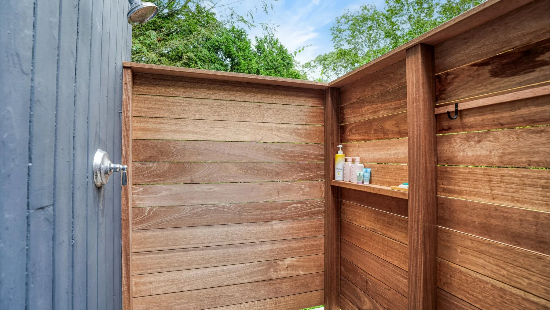 An outdoor shower area enclosed by wooden panels, with a showerhead and toiletries on a shelf.