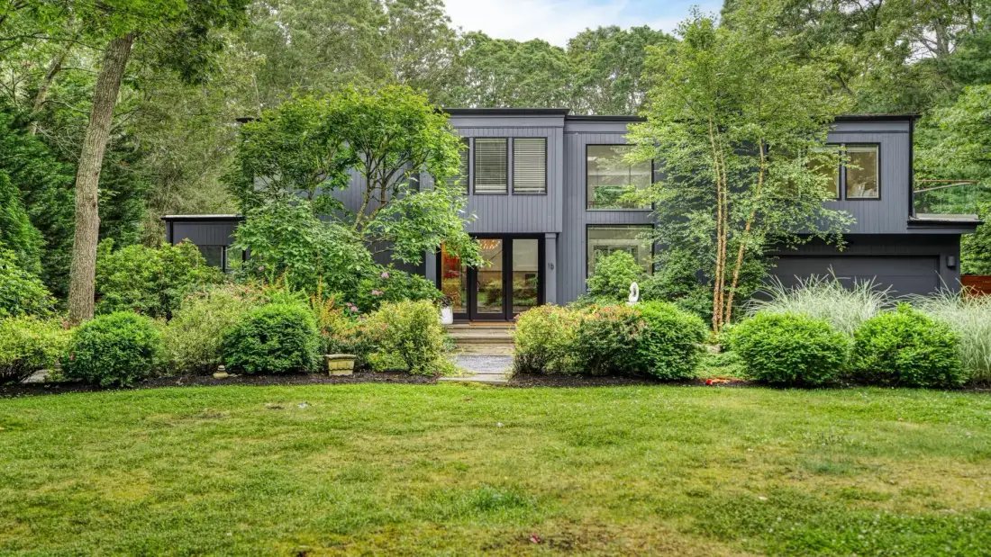 A modern house stands behind a lawn and dense green landscaping.