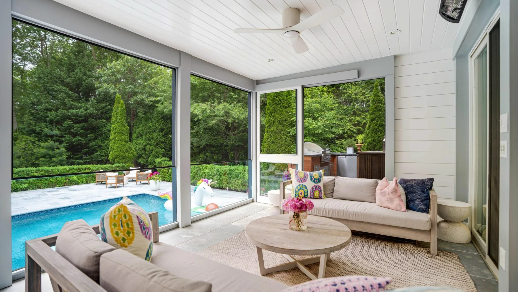 A bright sunroom with seating and large windows overlooking a pool and greenery.