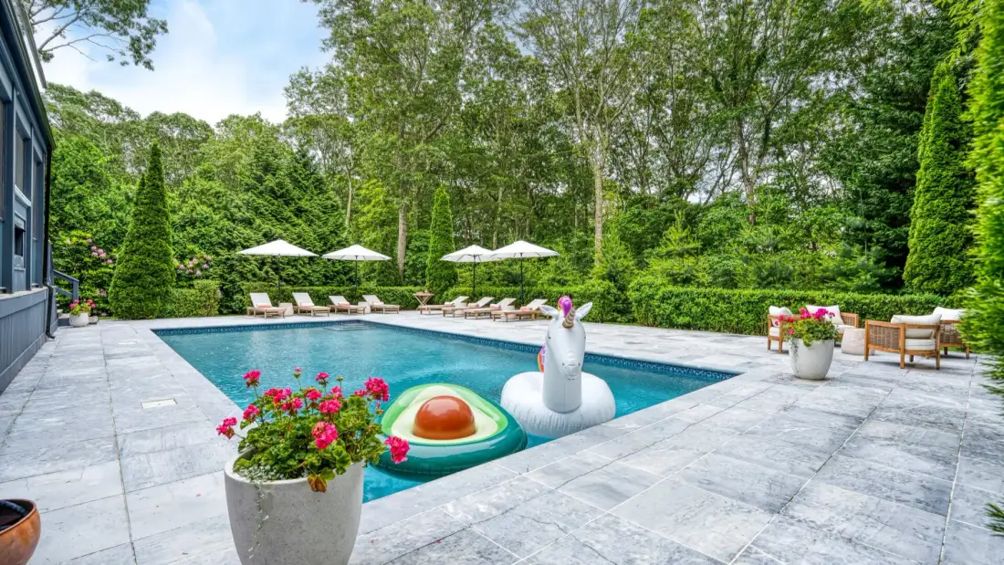 Una piscina ocupa un patio pavimentado rodeado de árboles y plantas con flores.