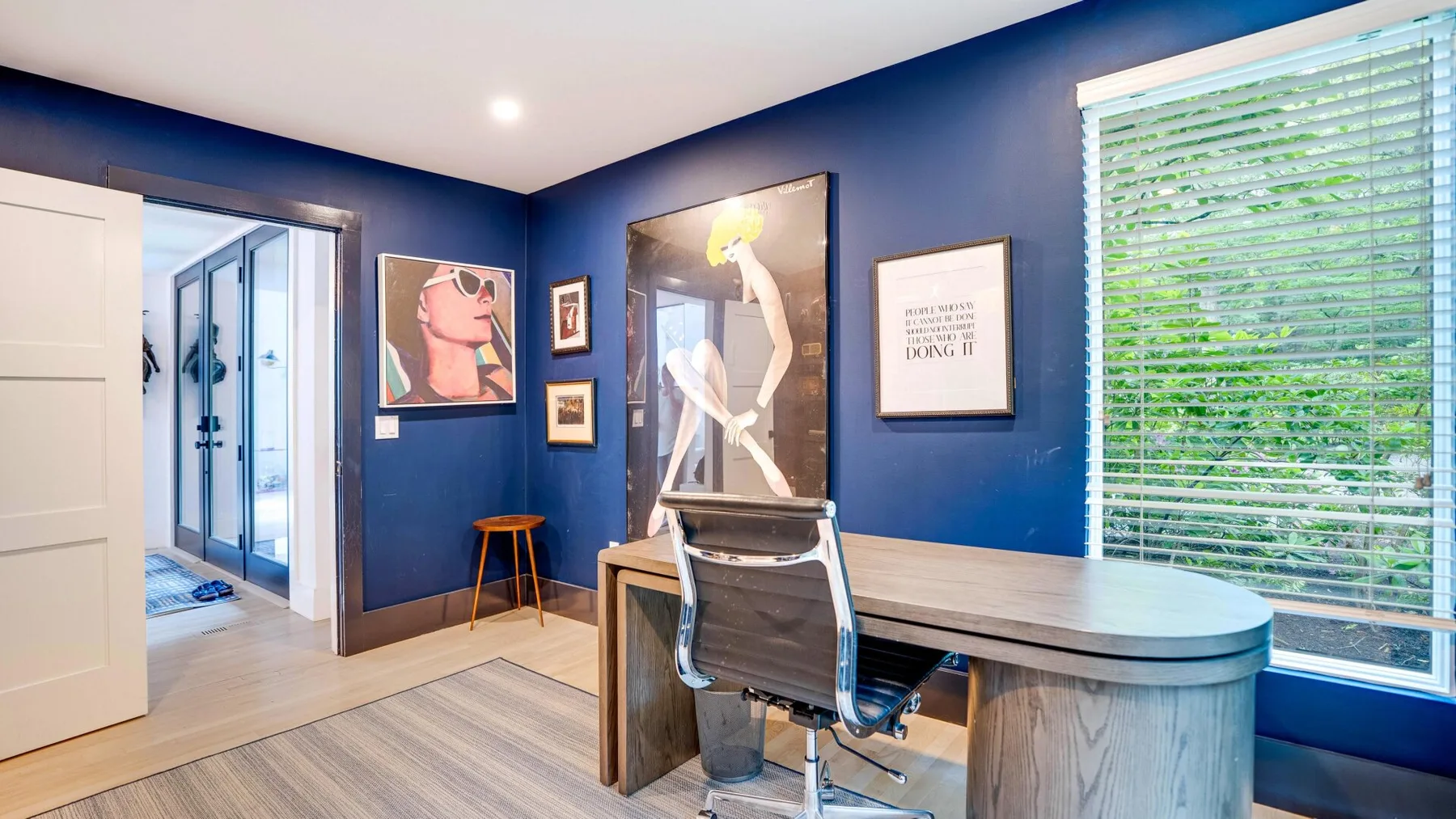 A blue-walled room with a desk, office chair, framed artwork, and a large window.