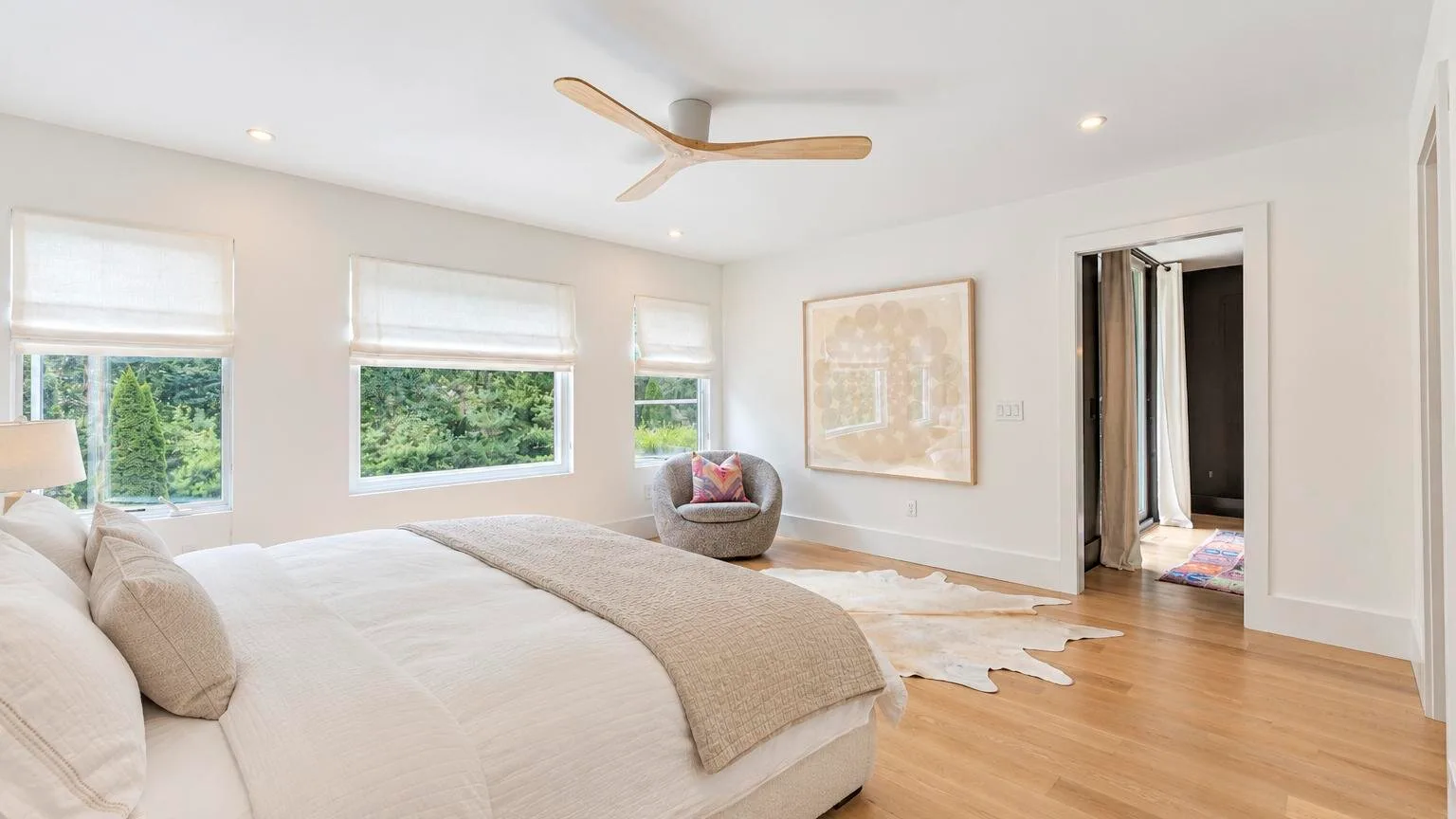 A bright bedroom with a large bed, a chair, and windows overlooking greenery.