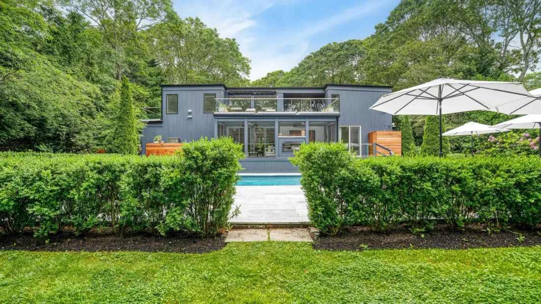 A modern house stands behind a hedge, with a swimming pool in front.
