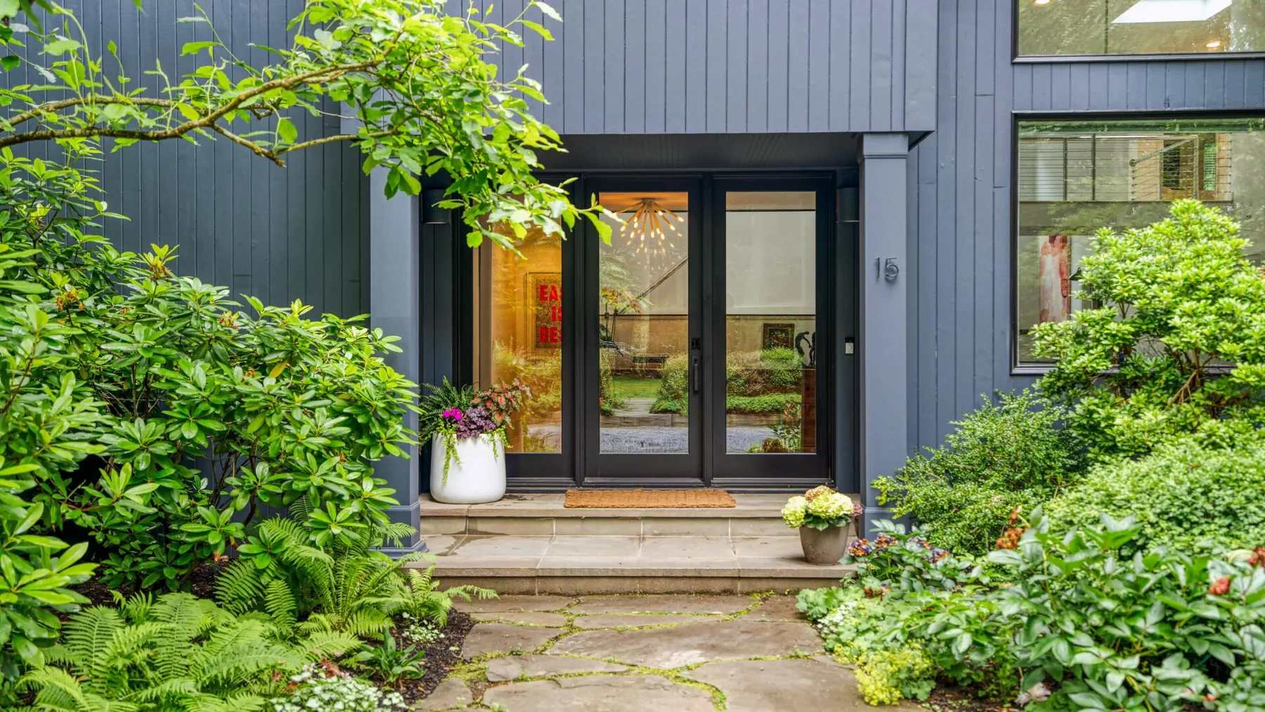 A modern home entrance opens onto a stone path bordered by leafy plants.