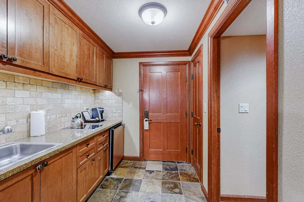 Una cocina estrecha con gabinetes de madera y piso de baldosas conduce a una puerta cerrada.